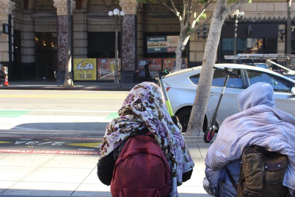 Two homeless individuals sit on 7th Street in downtown Los Angeles as a nearby restaurant promotes Super Bowl 56 at SoFi Stadium.