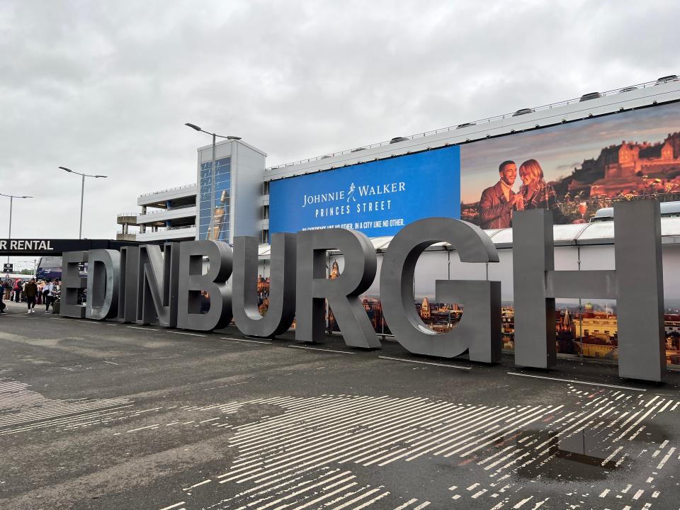 A large sign reads Edinburgh in gray letters in front of a Johnnie Walker billboard at Edinburgh Airport.