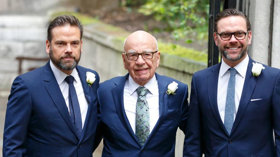 lachlan murdoch, rupert murdoph, and james murdoch pose for a photo while standing outside, the men wear matching navy suits with white shirts and various blue ties, each has a white rose on his lapel