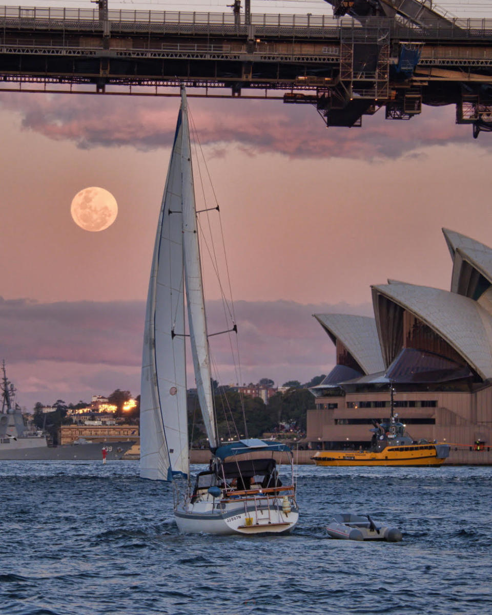 a full moon shines in a dusty pink sky above water. A small sailing boat is in the foreground of the image.