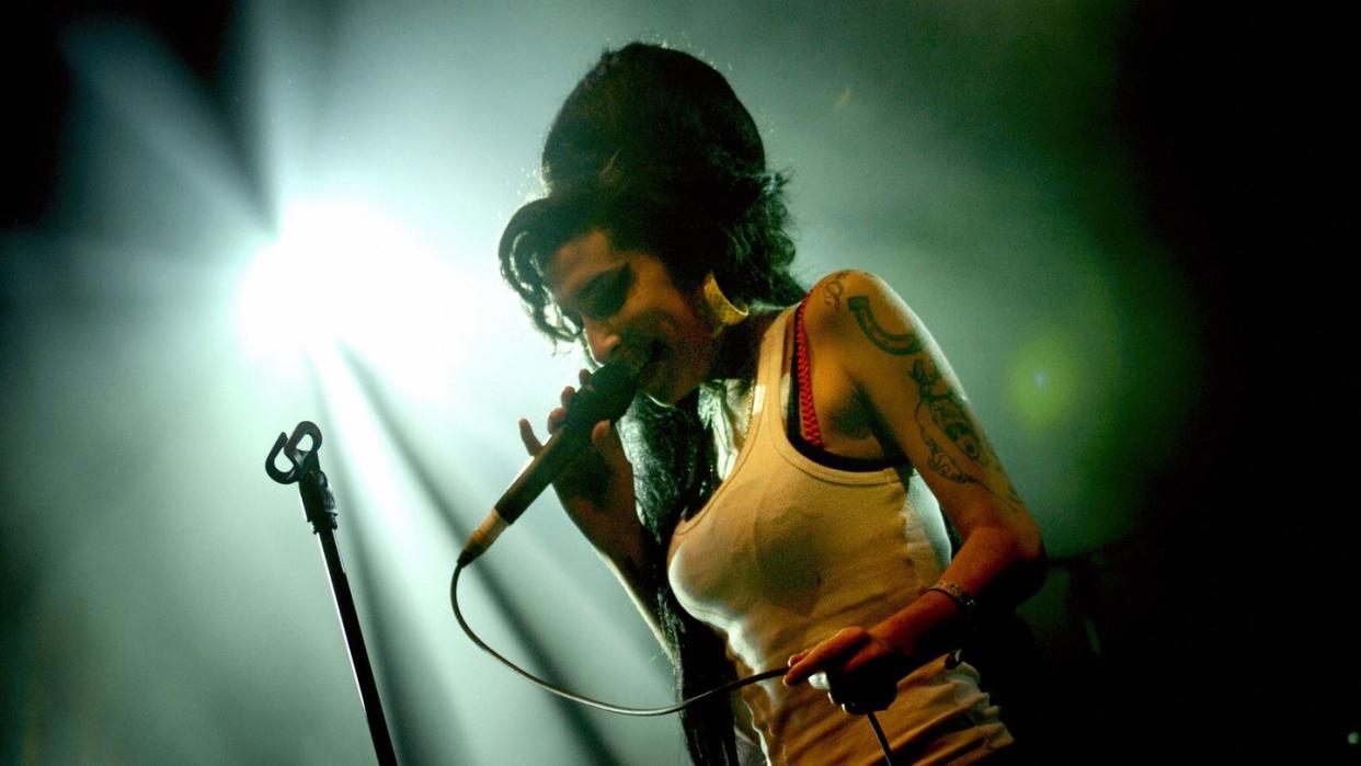 amy winehouse sings into a microphone she holds in one hand, she smiles and stands next to a microphone stand, she wears a white tank top