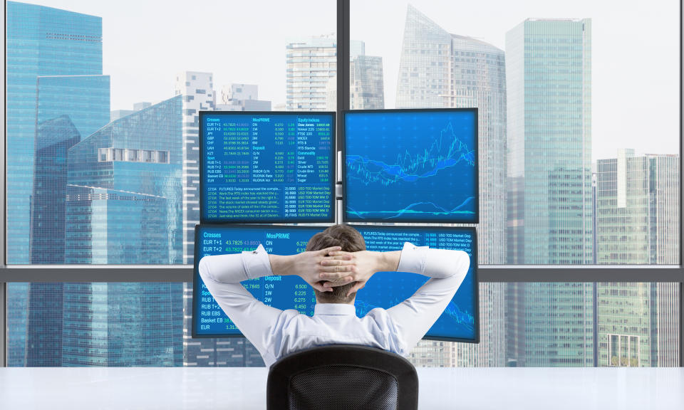 A man, seen from behind, sits with his hands clasped behind his back, as he looks at four computer screens full of data. Behind the computer screens is a window looking out on a city skyline.