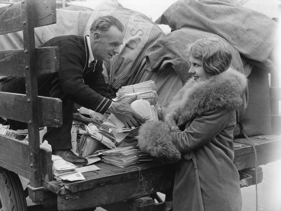 A postal worker in a suit hands mail to a woman in a fur coat