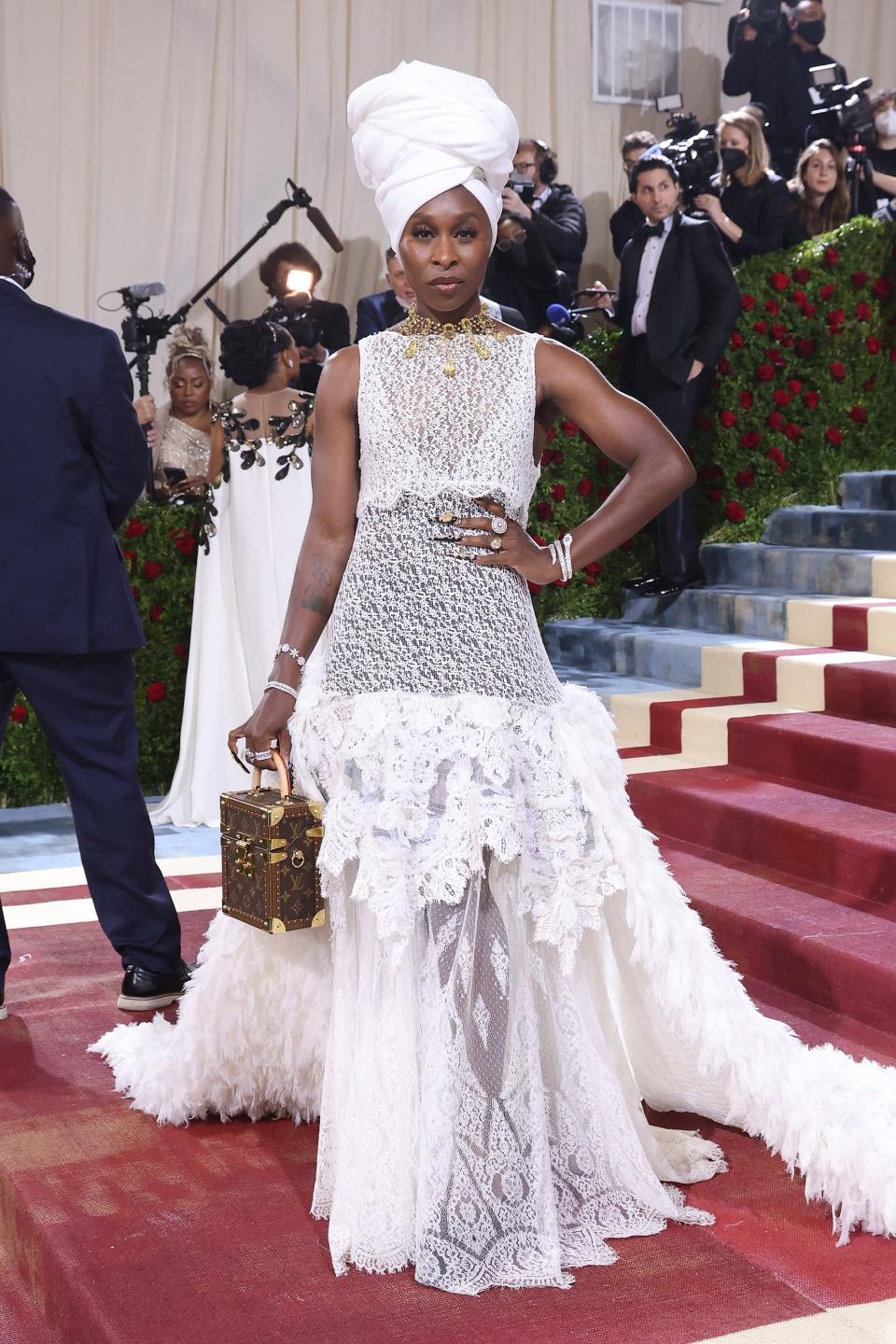 Cynthia in a large white wrapped headdress. She's wearing a shear white lace high neck dress that flares into ruffles and a feather train at her hips. She's holding a small brown Louis Vuitton boxy bag.