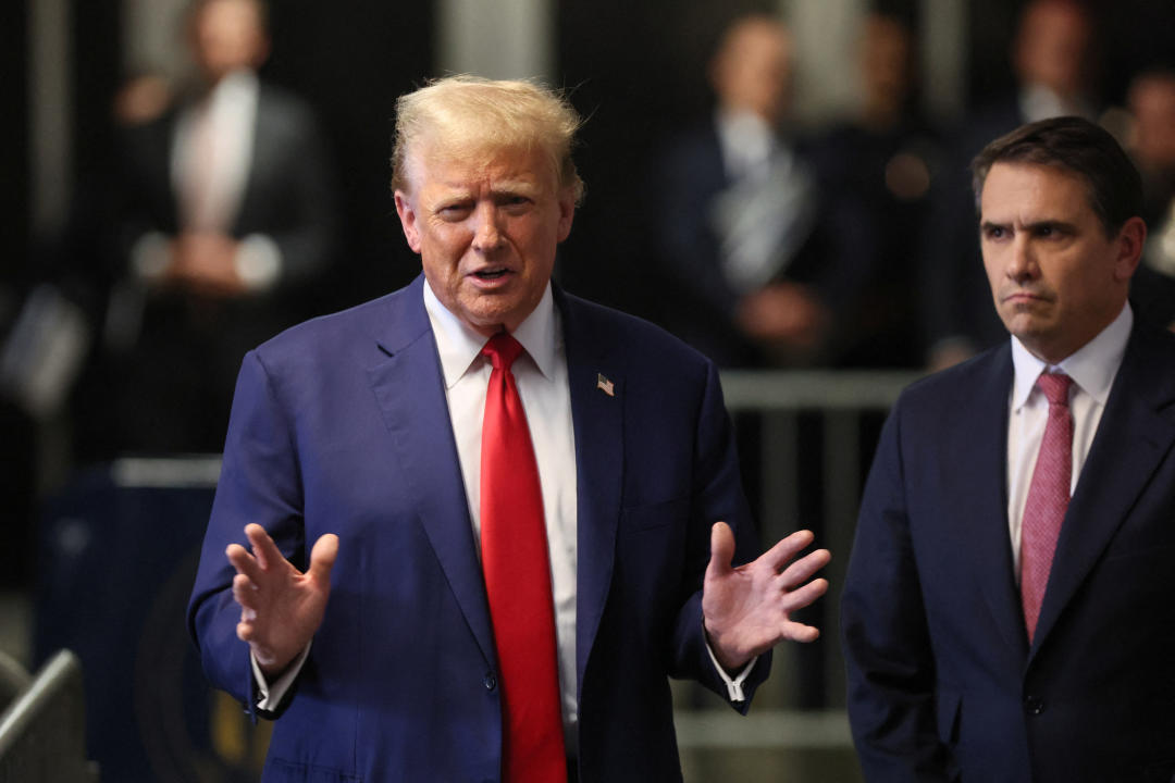 Donald Trump, in signature red tie and gesticulating, speaks to the media with his attorney Todd Blanche at his left.