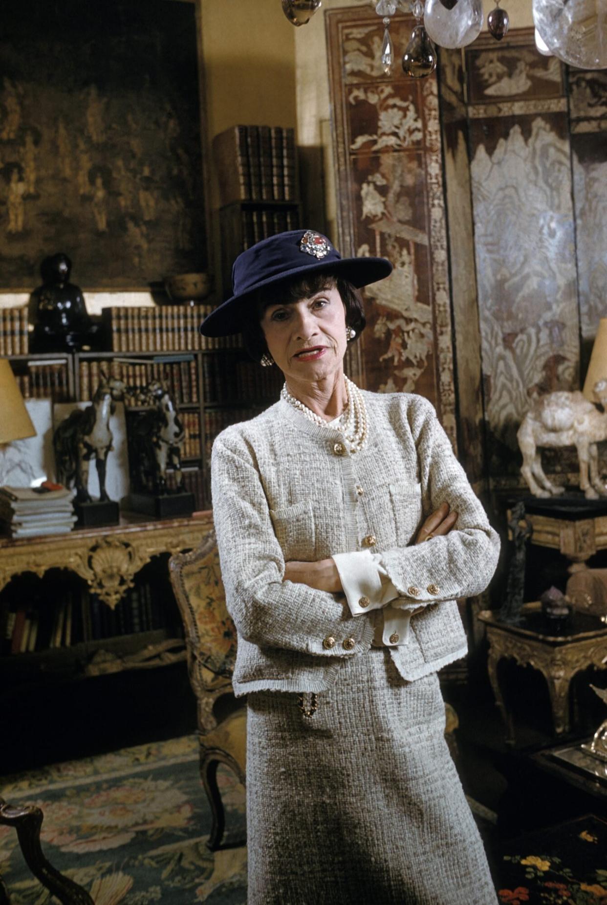 Coco Chanel wearing her signature tweed suit in cream with a pearl necklace and navy hat in her later years