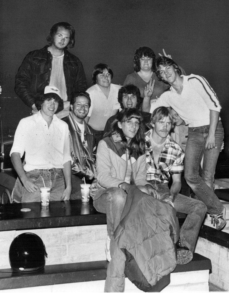 Group of 9 people posing casually with playful gestures, some holding cups, in a low-lit setting