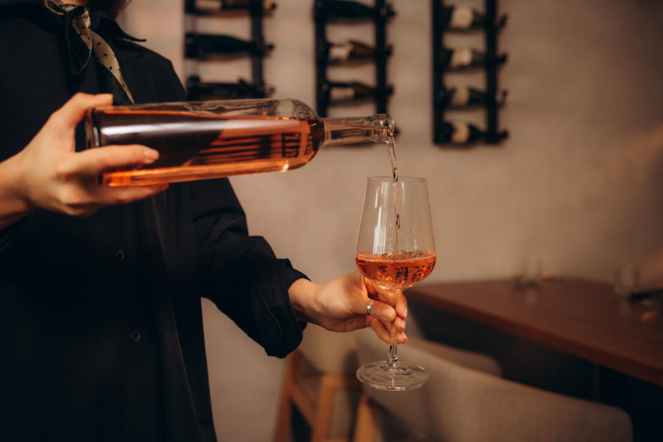 A person pours rose wine from a bottle into a glass in a wine-tasting room with wine bottles on the wall behind them