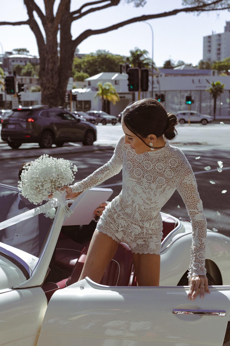 A woman in a white, lace romper climbs into a convertible while holding a bouquet of flowers.