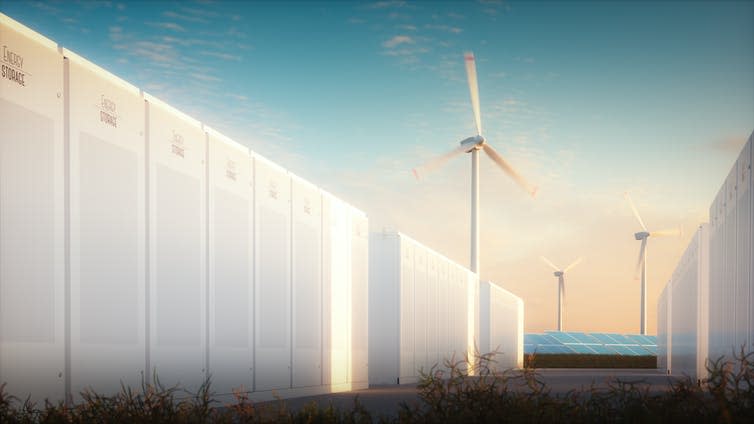 A wall of large, white batteries with wind turbines and solar panels in the background.