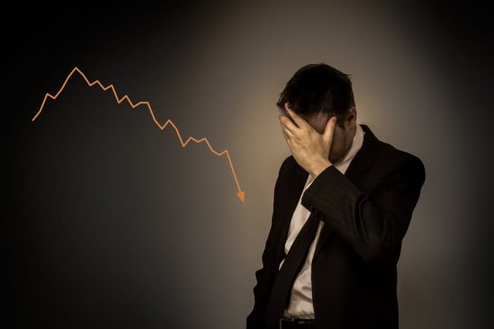Man in suit with hand over his face standing next to a chart that's trending downward.
