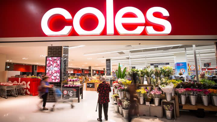 Photo shows the entryway to a Coles supermarket including a display of fresh flowers for sale, with an elderly woman standing in front of it.