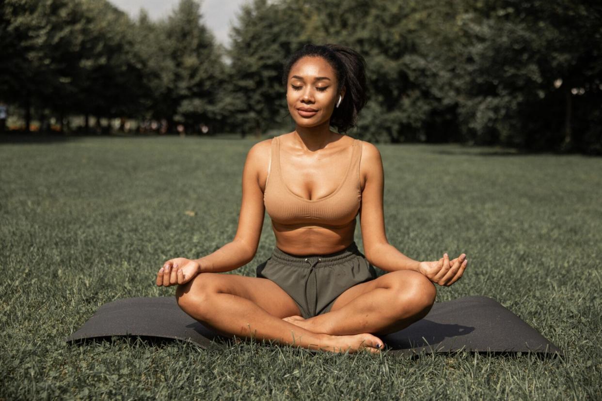 Black woman sitting cross legged on a yoga mat in a park relaxed