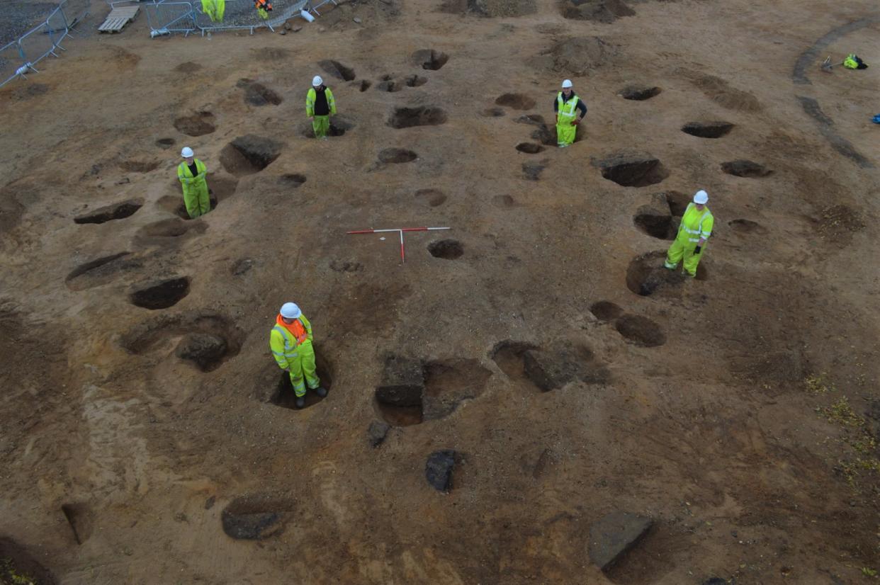 Five archaeologists in hi-vis clothing standing in postholes of a prehistoric roundhouse