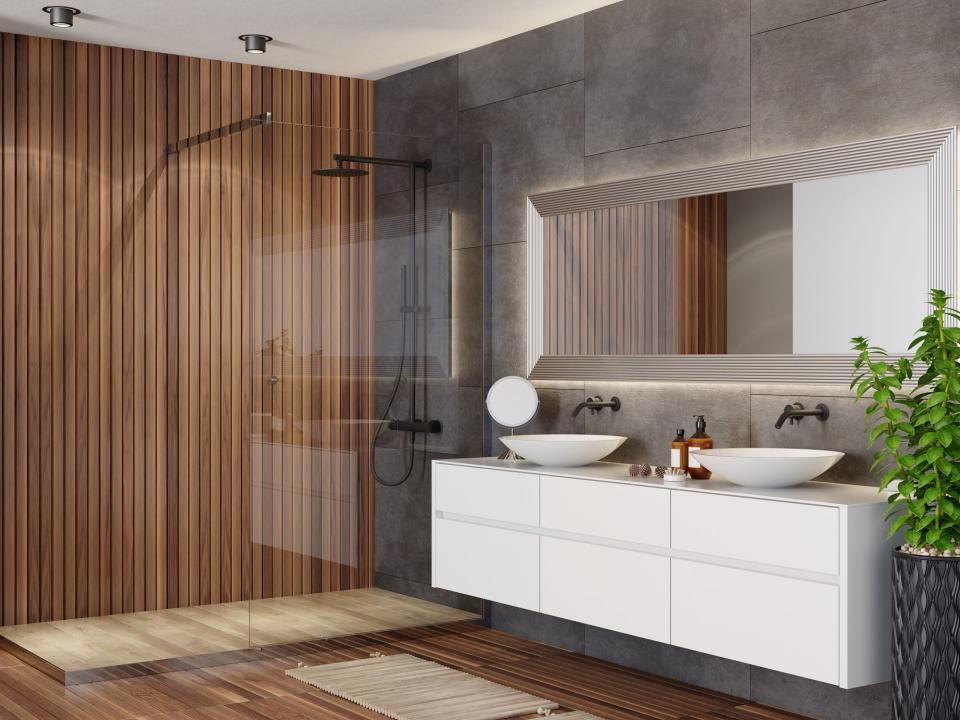 A bathroom with a white floating sink and two faucets, a large mirror, a gray stone wall, and a shower with a clear-glass door and a wooden paneled shower wall