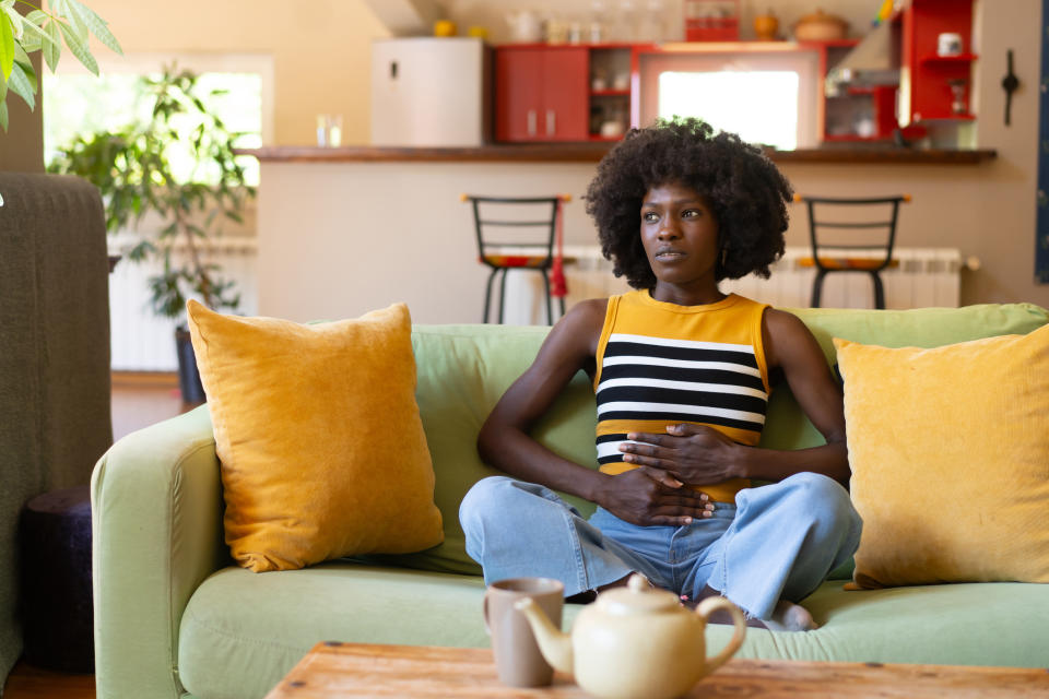 A person sits on a sofa in a kitchen. They appear to be experiencing stomach discomfort, holding their abdomen. A teapot and cups are on the table in front