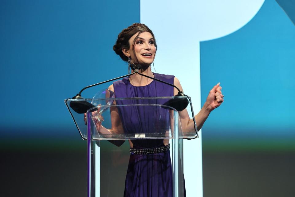dylan mulvaney, wearing a purple dress, standing in front of a transparent podium with two microphones and speaking