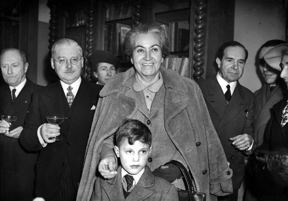 gabriela mistral smiles and looks right of the camera, she stands in a room with over people and wears a coat over her suit jacket and collared blouse