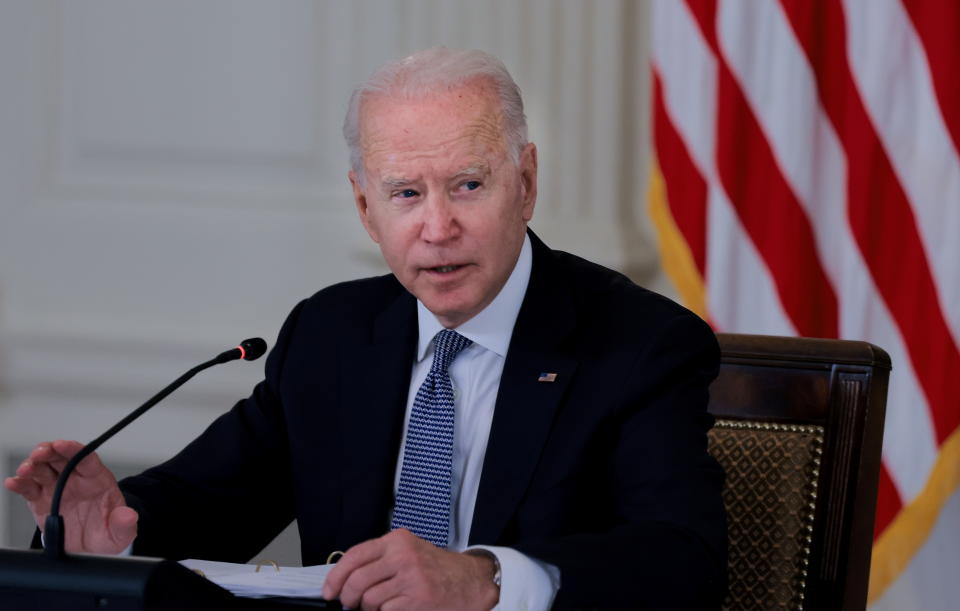 President Biden sits at a desk in front of an American flag and leans toward a single microphone.