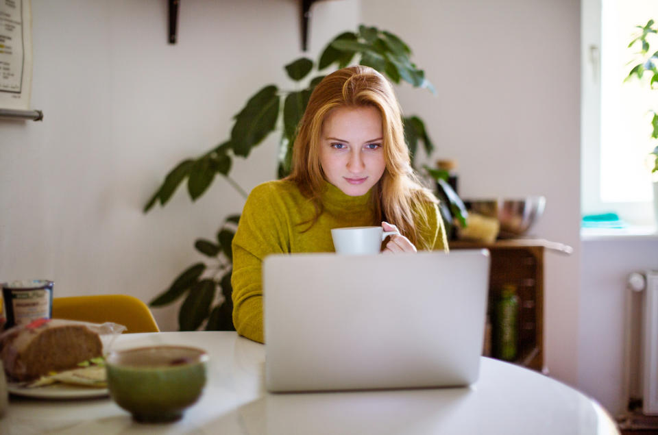 A woman with long hair sits at a kitchen table, holding a mug and looking at a laptop screen. A plant is in the background