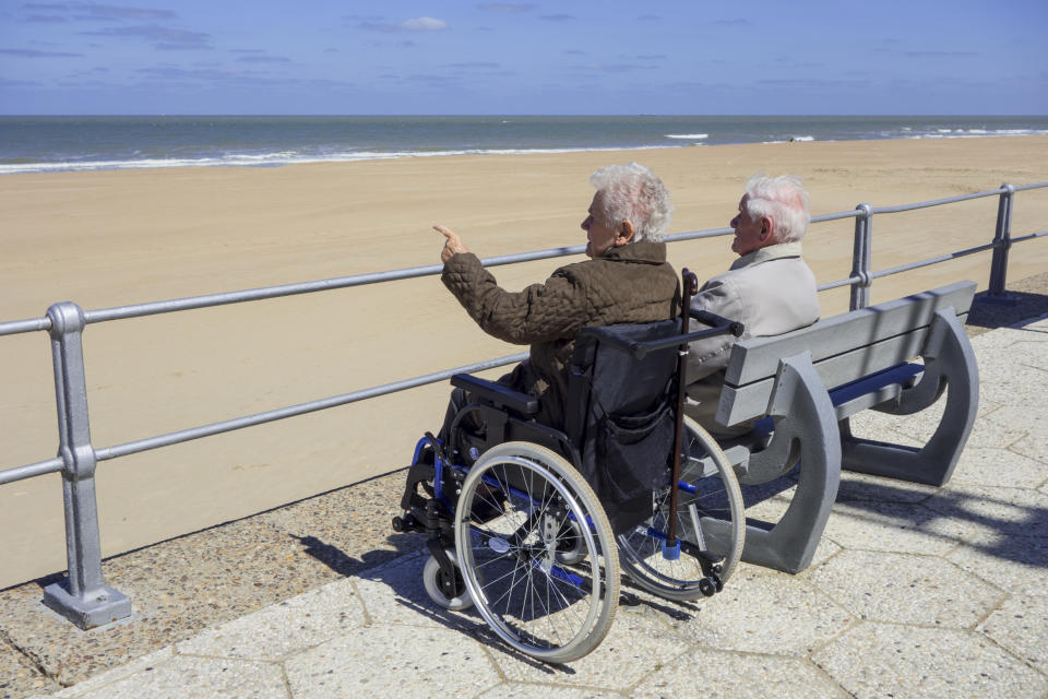 An elderly woman in a wheelchair and her retired husband sit on a bench at a promenade along the coast on a cold sunny day in spring.