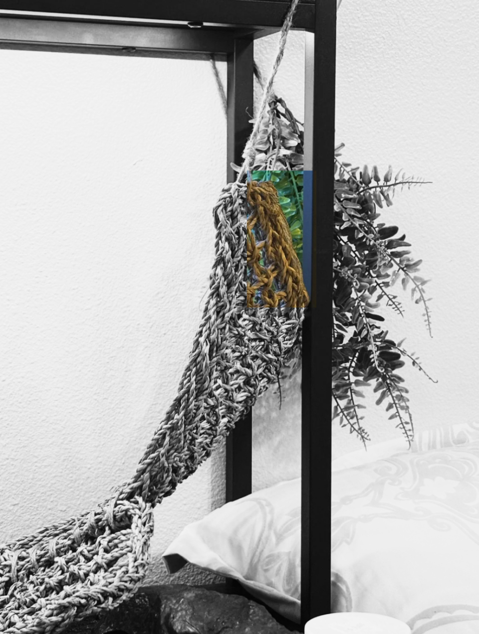 A small knitted hammock hangs from a metal frame next to a bed, with a green leafy plant behind it