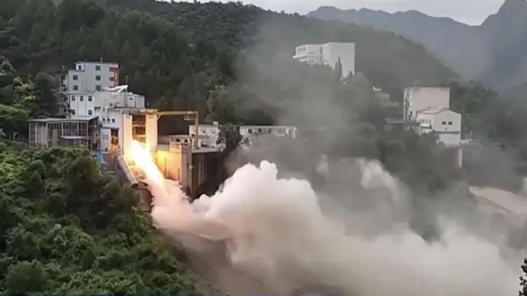  A powerful fire shoots downhill from the opening of a building nestled into the side of a green, hilly mountainous slope. Smoke fills the right side area 