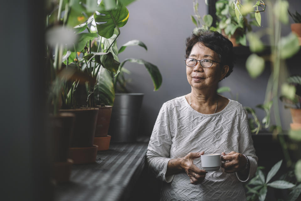 An elderly woman with short hair and glasses stands among potted plants, holding a teacup, wearing a light patterned top