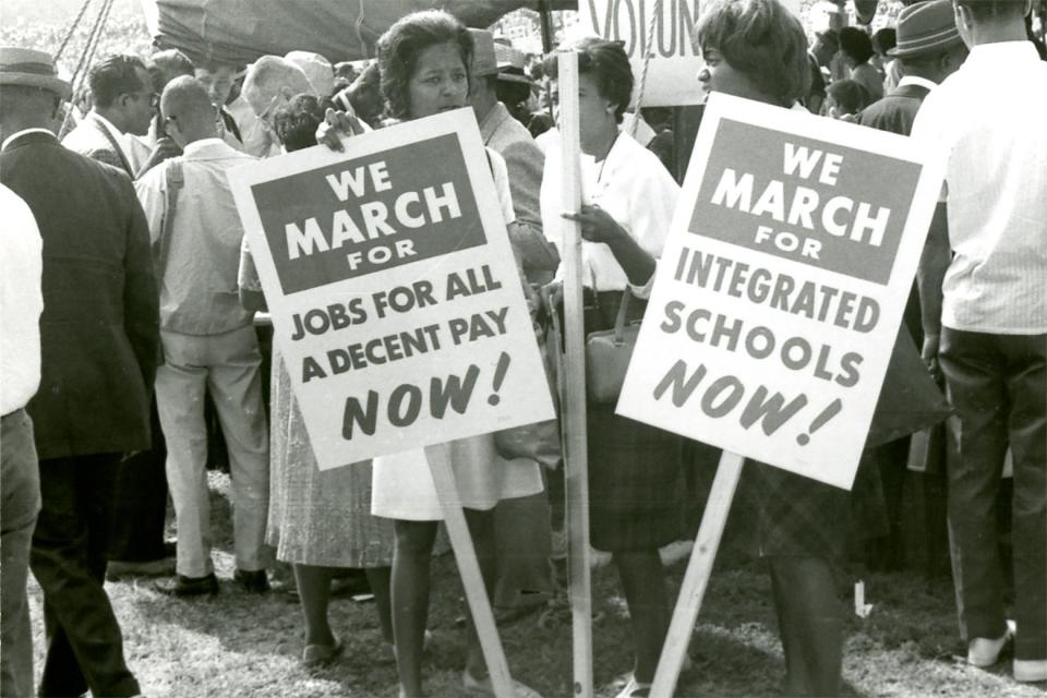 Protestors at the 1963 march hold signs that say "We march for integrated schools NOW!"