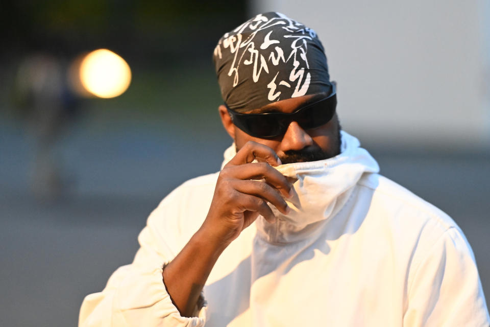 Kanye West in a casual white jacket, black bandana with white designs, and dark sunglasses, holding the jacket's collar near his face