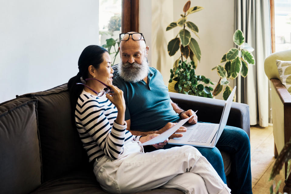 Senior interracial couple using a laptop and going through paperwork to pay bills online on the couch at home. Mature husband and wife talking and planning their retirement on a pc on the sofa