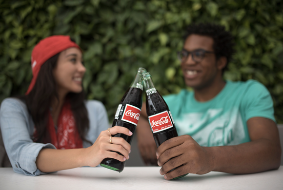 A young woman and young man giving cheers to each other while holding bottles of Coke.