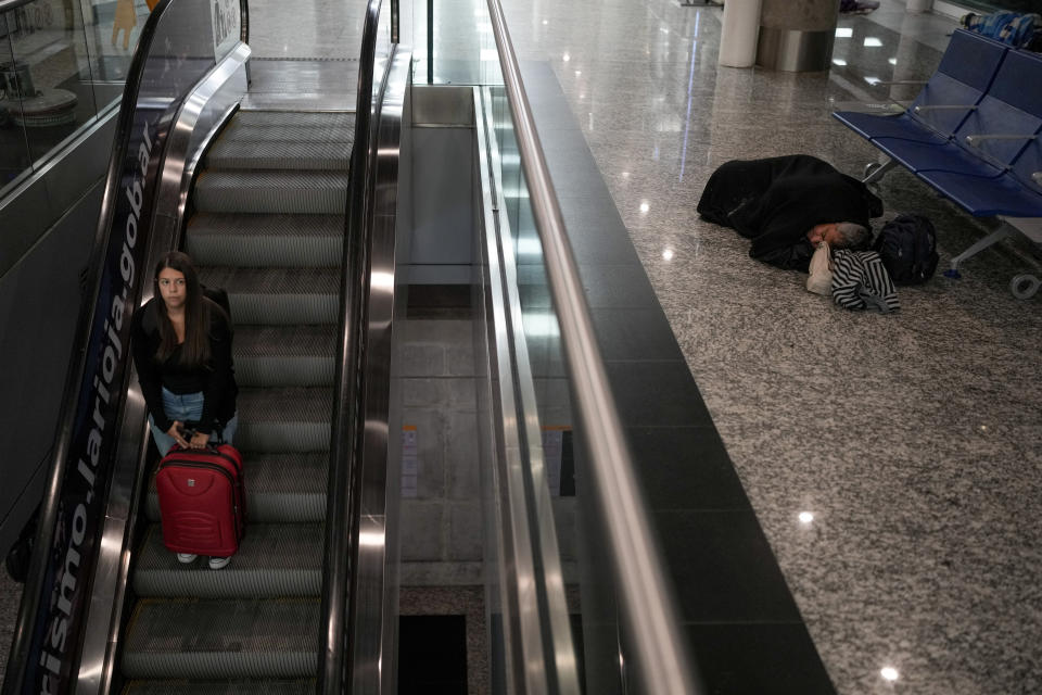 A passenger descends an escalator as a homeless man sleeps near a row of seats at the at the Jorge Newbery international airport, commonly known as Aeroparque, in Buenos Aires, Argentina, Thursday, April 6, 2023. Despite the bustling activity of passengers, over 100 homeless people sleep at Aeroparque each night. (AP Photo/Natacha Pisarenko)