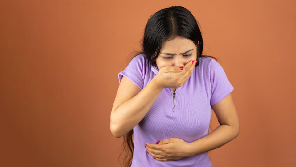 A photo of a woman in a purple t-shirt clutching her stomach and mouth as though she is about to be sick