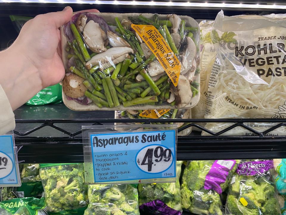 hand holding a package of asparagus saute in front of the preprepared veggies fridge at trader joes