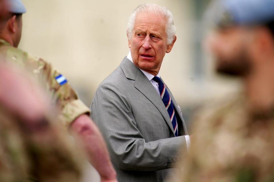 King Charles III wearing a grey suit and a striped blue tie standing near British army personnel.