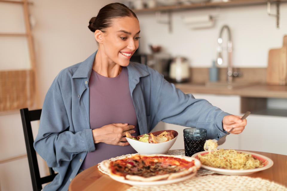 A woman sits at a table with various dishes including pizza, pasta, and salad in front of her. She is smiling and holding a fork, ready to eat