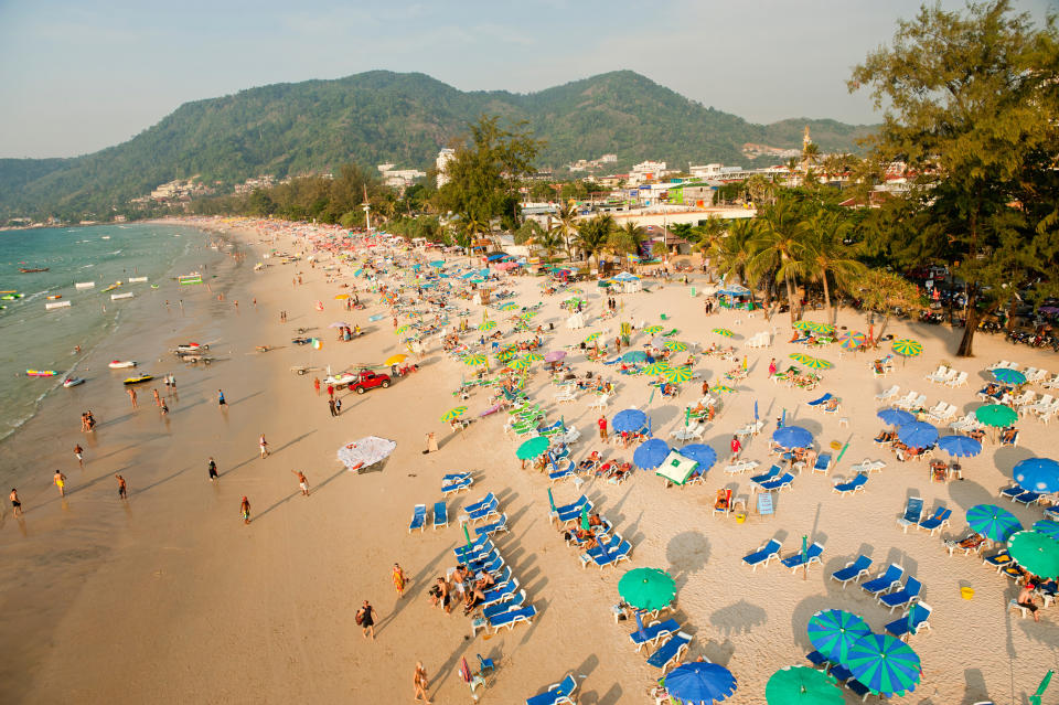 A busy tropical beach with numerous people sunbathing and swimming. The shoreline is lined with multi-colored umbrellas and lounge chairs. Green hills and buildings are in the background