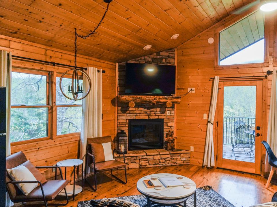 Inside the cabin's living room, from left to right, there's a tiny table with two chairs and a spherical chandelier  hanging in front of a window; a stack of two white coffee tables that are circular, a stone fireplace with a TV mounted above it, and a windowed door to the balcony with another window above it shaped like a trapezoid.