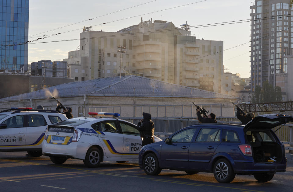 Beside two official cars with blue-and-yellow Ukrainian markings, parked on a road not far from two tower blocks, three marksmen fire into the air.