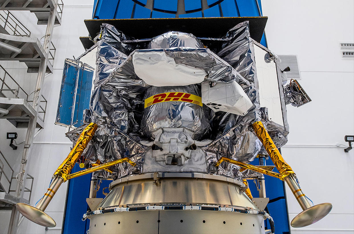  A spacecraft wrapped in silver foil sits in a white-walled room. 