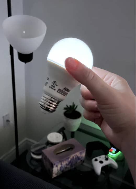 A hand holding a lit LED light bulb in a living room setup, including a floor lamp, small table with a tissue box, and a game controller