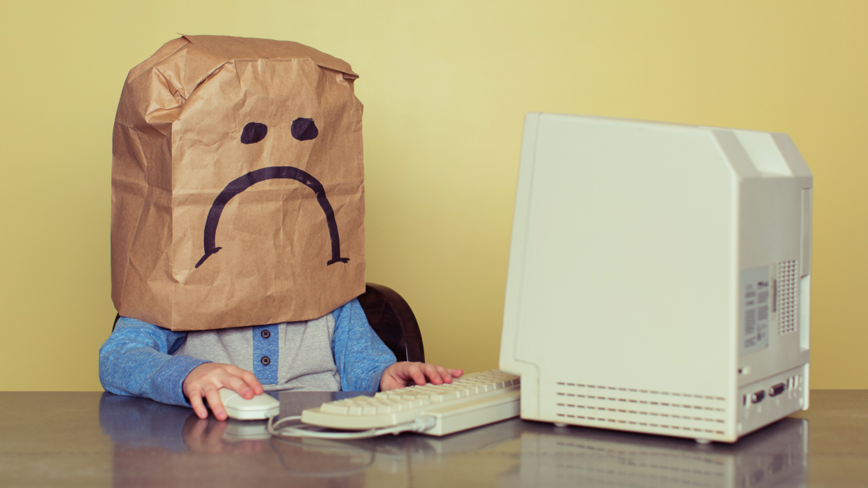  A child with a sad paper bag on their head sits staring at an old PC. 