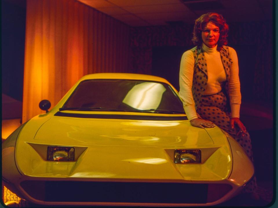 A vintage photo of a woman leaning onto a car prototype in a showroom.