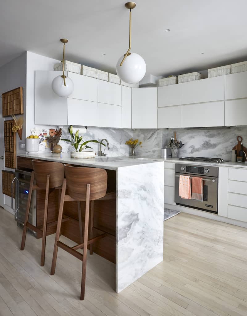 Kitchen with marble bar/counter/backsplash, curve-backed wooden stools, white cabinets, fridge, white globe pendant lights