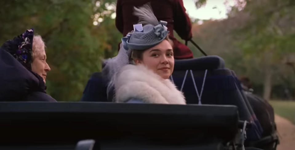 Florence Pugh as Amy March, wearing period clothing and a bonnet sits in a horse-drawn carriage, looking back over her shoulder with a gentle smile as an older woman sits beside her