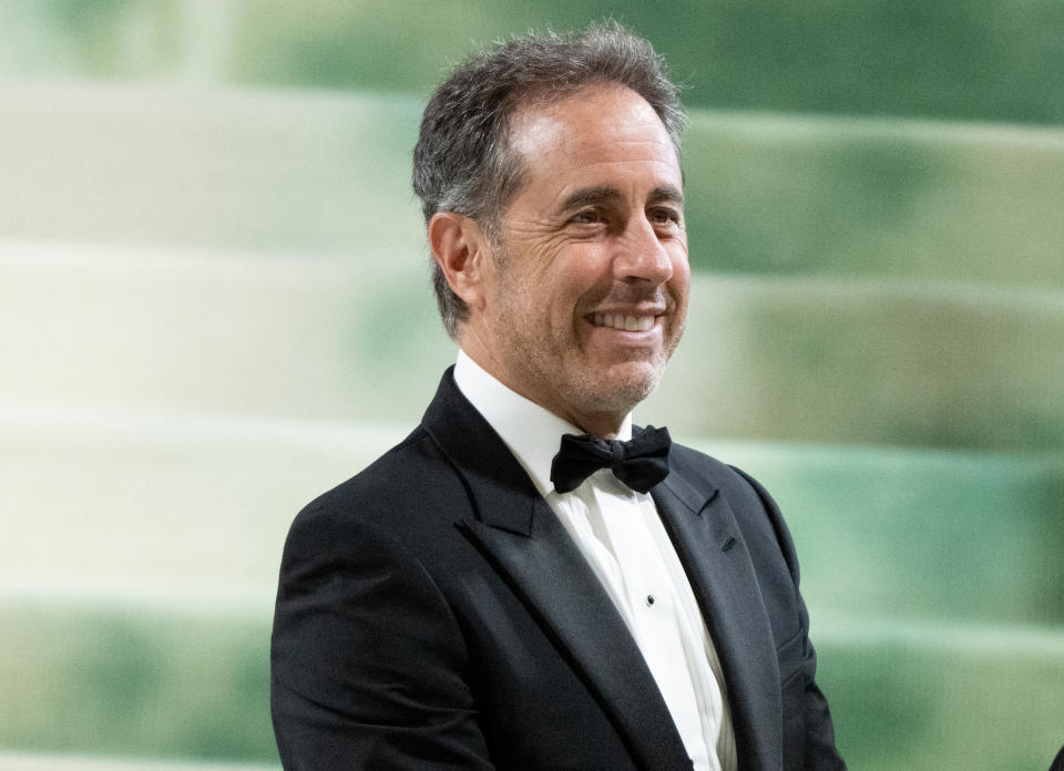 Jerry Seinfeld wearing a black tuxedo with a white shirt and black bow tie, smiling on the red carpet