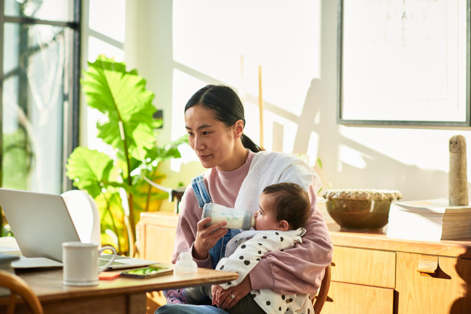 Adult feeding a baby while working at a laptop in a home environment