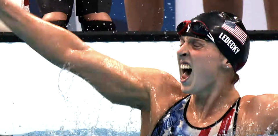 Katie Ledecky celebrates in pool after winning a race, wearing a swim cap with the American flag and "Ledecky" printed on the side, and a swimsuit