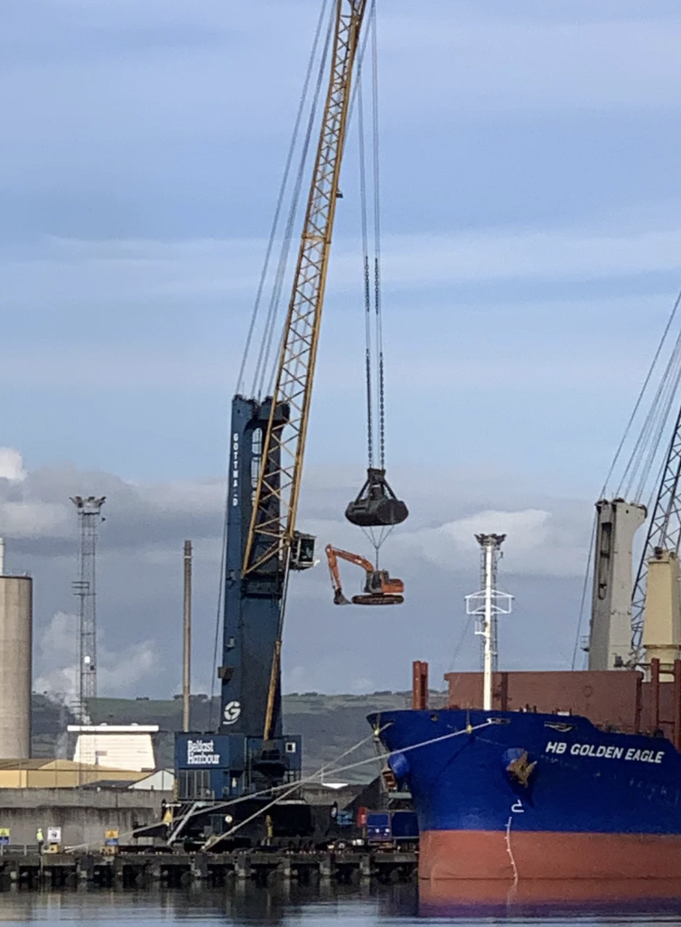 A large crane lifts an excavator onto a blue cargo ship named "HB Golden Eagle" at a harbor. Industrial facilities and other machinery are in the background